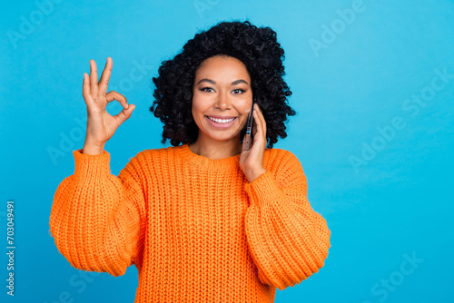 Photo portrait of young woman talk phone show okey symbol dressed stylish knitted orange clothes isolated on blue color background