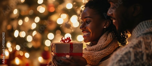 Blissful woman giving a gift with spouse and holiday decor in the background.