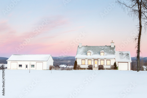 Sunrise winter view of steep shingled roofed patrimonial white wooden house and barn with Laurentian mountains in the background, St. Pierre, Island of Orleans, Quebec