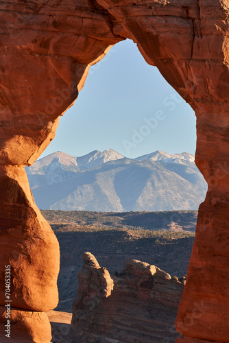 The La Sal mountain range perfectly framed by Delicate Arch in Arches National Park. A small amount of snow is on the top of the peaks. Sunlight bathes them from an angle.