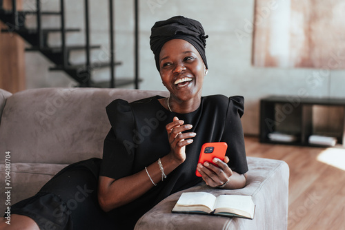 Radiant woman in black headscarf and dress enjoying a moment with her red smartphone. Happy African girl in turban working home.