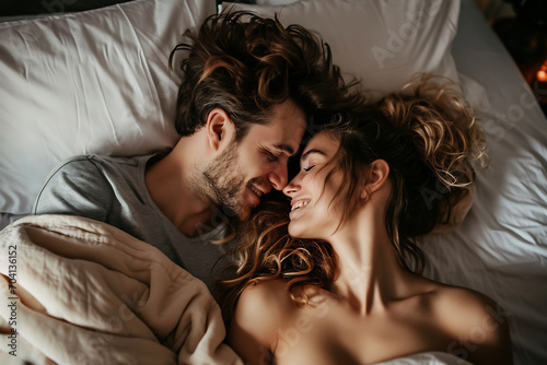 Young beautiful and loving couple wake up while lying on white pillows under the covers on soft white bed at the morning