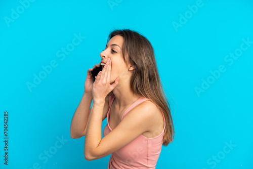 Young caucasian woman using mobile phone isolated on blue background shouting with mouth wide open to the side