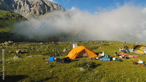 A campsite for overnight camping in the mountains. Orange tent, rucksacks and sleeping bags lying on the grass. Rocky mountains and fog in the background.