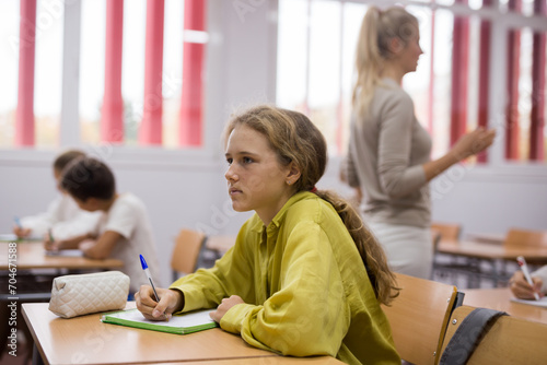 Focused school girl sitting at school desk in classroom on background with classmates