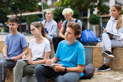 Attentive students write lesson in a notebook sitting on a stone street parapet