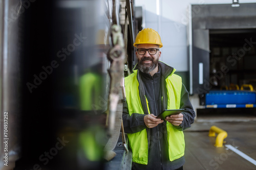 Portrait of smiling warehouse receiver standing by blue truck and holding tablet. Receiving clerk looking at cargo details, checking delivered items or goods against order.