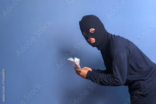Bank robber wearing balaclava holding bunch of stollen dollars, isolated on blue studio background