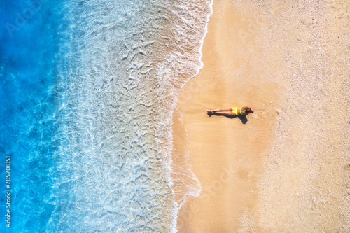 Aerial view of the beautiful young lying woman on the tropical sandy beach near sea with waves at sunset. Summer vacation in Lefkada island, Greece. Top view of slim girl, clear azure water. Seaside
