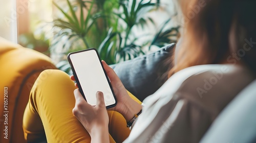 Happy young woman holding mobile smartphone with blank white screen background while resting on the sofa in living room at home. Watching movies on the phone