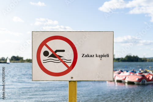 No Swimming sign post plaque, with water in background. No swim area signage pillar, mounted on the shore of lake in Poland. Text in Polish Zakaz kąpieli, means Swimming prohibited.