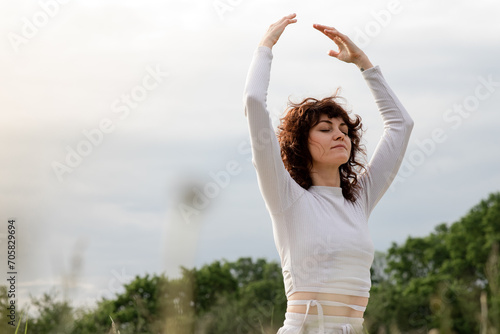 Concept of morning relaxation on nature background. Athlete girl raised arms up sitting yoga mat. Sports woman fitness coach active breathing fresh air. Stretching exercise in nature