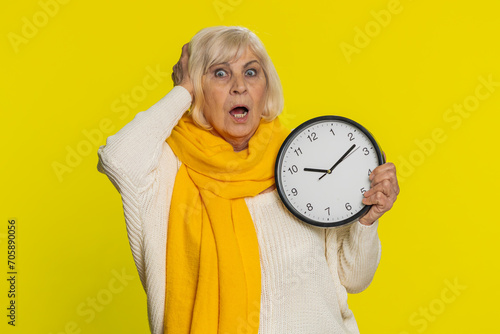 Senior old woman with anxiety checking time on clock, running late to work being in delay, deadline. Mature grandmother looking at hour, minutes, worrying to be punctual isolated on yellow background