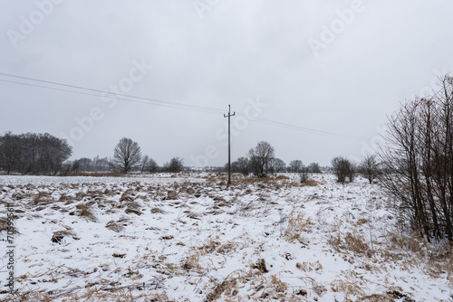 Ścieżka elektryczna idąca przez zaśnieżone pole porośnięte krzakami w popołudniowych godzinach pochmurnego szarego dnia w Zachodniej Polsce