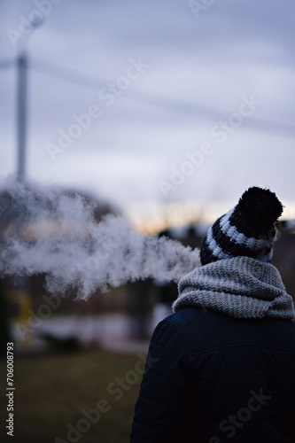 Postać osoby tyłem, w zimowej czapce i szaliku, wydmuchującej chmurę dymu na tle zimowego pochmurnego nieba