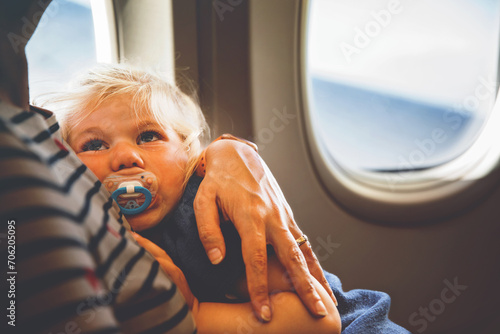 Woman traveling with little child by airplane. Sad tired toddler girl sitting with mum by aircraft window. Motherhood concept. Crying baby.