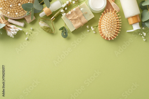 Organic self-care essentials. Top view of jade roller, skincare products, wooden hairbrush, cellulite brush, artisanal soap, floss, cotton buds, eucalyptus, gypsophila on green backdrop, space for ad