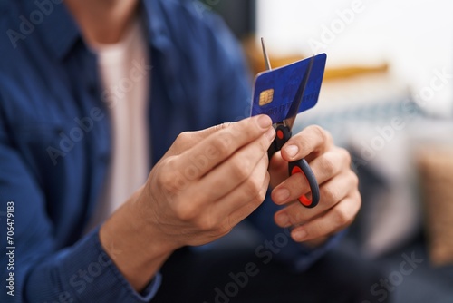 Young caucasian man cutting credit card sitting on sofa at home