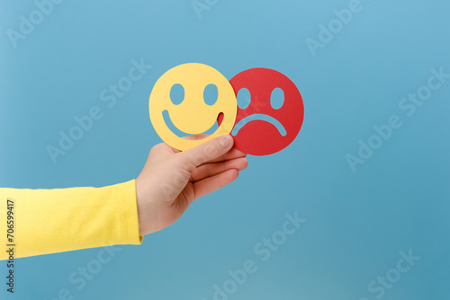 Close up of female hand holding happy and angry face paper, isolated on blue studio background. Feedback rating, emotional intelligence, control, mental health assessment, bipolar disorder concept