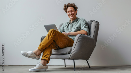 Young Caucasian man with curly hair, comfortably seated in a modern gray armchair, smiling and relaxed while using a laptop on his lap