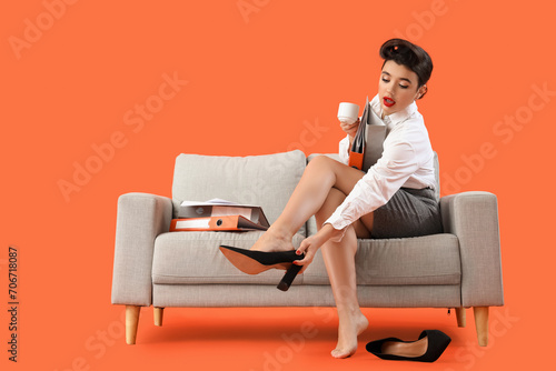 Young pin-up businesswoman with coffee and folders taking off heels on sofa against orange background