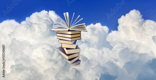 stack of books with an open book lying on top of the stack against a background of white clouds