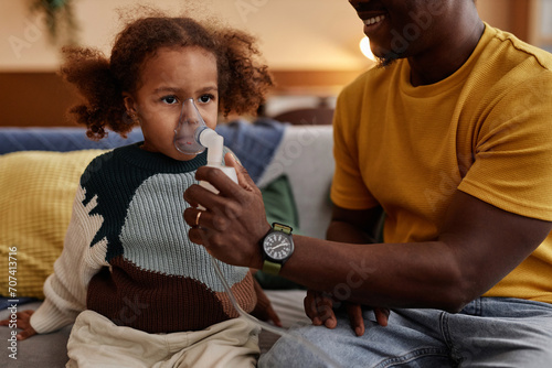 Cropped shot of smiling father putting nebulizer mask on face of unwell African American girl child while sitting on couch at home