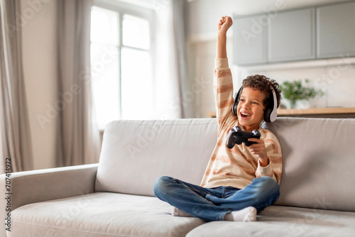 Little black boy in headphones holding joystick and raising hand in victory