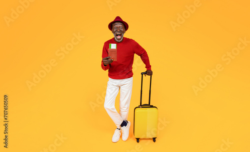 Happy excited senior african american man tourist in hat with yellow suitcase and open mouth show passport
