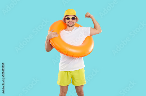 Cheerful man with inflatable swimming circle shows biceps on light blue background. Smiling male vacationer, humorous strong swimmer or beach lifeguard in T-shirt, shorts, panama and sunglasses.