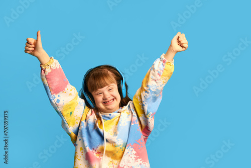 Happy smiling teen girl with down syndrome listening to music in headphones, expressing joy against blue background. Concept of acceptance, care, inclusion, health, diversity, emotions, equality
