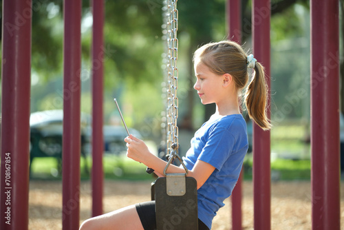 Young happy smiling teenager girl reading study homework on digital tablet during summer vacations sitting on swing in park