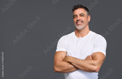 Happy fit sporty older man coach, middle aged sportsman athlete or personal trainer wearing white t-shirt showing muscles standing isolated on gray background advertising gym membership. Portrait.