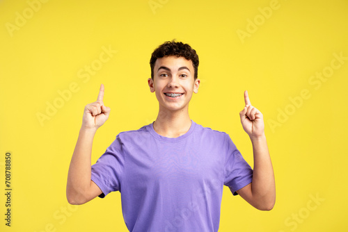 Handsome teenage boy with dental braces wearing purple t shirt pointing finger at copy space