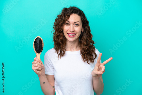 Young caucasian woman holding hairbrush isolated on blue background smiling and showing victory sign