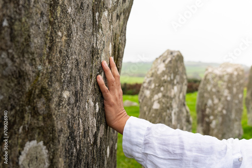 Close up of woman hand touching a large megalith belonging to the Drombeg Neolithic circle in County Cork in Ireland with other megaliths in the background and green meadows.