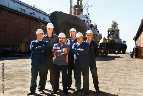 Maintenance crew poses in front of large vessel at shipyard dry dock. Engineers in hardhats, coveralls stand ready for maritime repairs. Industrial workers, docked ship, outdoor teamwork setting.