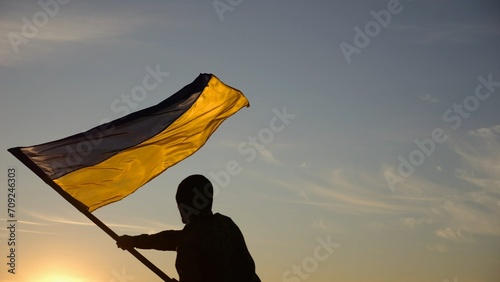Young man in military uniform waving flag of Ukraine against beautiful sunset at background. Male ukrainian army soldier lifted national banner at countryside. Victory against aggression