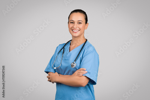 Happy nurse lady with folded arms posing wearing blue workwear and standing on gray studio background, smiling to camera