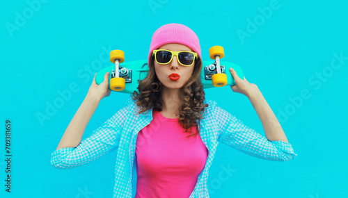 Portrait of stylish modern young woman with skateboard posing in colorful vivid clothes, pink hat on blue studio background
