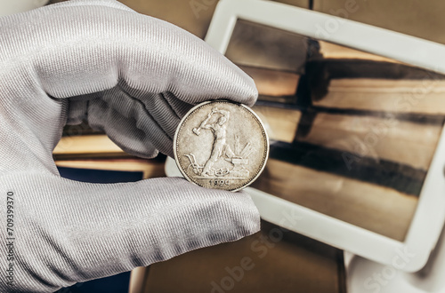 Photo of a person's male hand in white gloves holding a soviet silver 1924 half ruble coin on stack of books and magnifying glass background. Numismatics hobby concept.