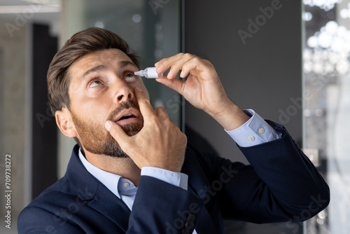 Close-up photo of a young businessman sitting in the office at a desk in a suit and instilling medicine into his eye, treating inflammation, pain, chronic fatigue