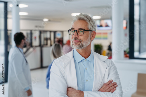 Portrait of confident mature doctor standing in Hospital corridor. Handsome doctor with gray hair wearing white coat, stethoscope around neck standing in modern private clinic, looking at camera.