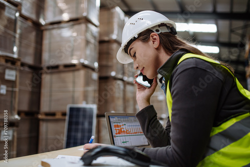 Warehouse worker checking delivery, stock in warehouse on computer, pc, while phone calling with contractor. Warehouse manager using warehouse management software, app.
