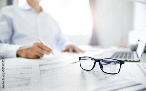 Close-up shot at eyeglasses, blurred businessman review many documents placed on the desk and using laptop meeting online with business group to plan and start new business