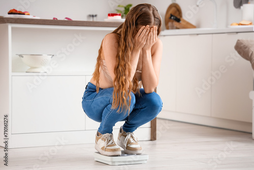 Upset young woman in tight jeans on scales at home. Weight gain concept