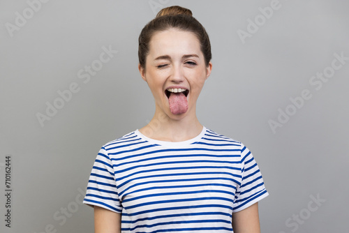 Portrait of disobedient crazy funny woman wearing striped T-shirt showing tongue out and closed her eyes displeased, behaving unruly naughty. Indoor studio shot isolated on gray background.