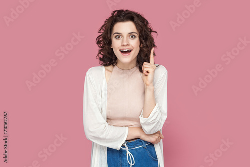 Portrait of clever smart happy woman with curly hair wearing casual style outfit raises her index finger up, having good idea. Indoor studio shot isolated on pink background.