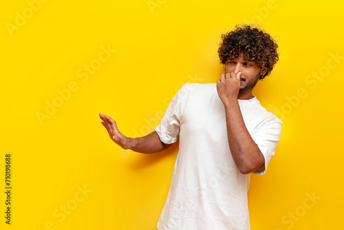 young curly indian man showing disgust and covering his nose with hand on yellow isolated background, guy refuses bad smell and rejects stench