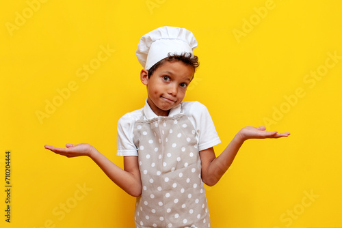 unsure african american boy in uniform and chef's hat shrugs his shoulders on yellow isolated background, confused teenager child in apron raises his hands and shows uncertainty